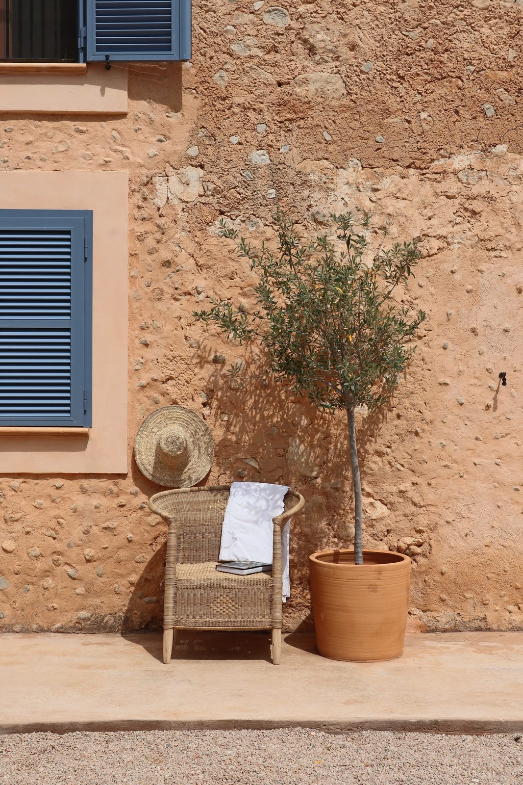 My African Med, a cane chair and straw hat outside a rustic Mallorca house with blue shutters and an olive tree in a terracotta pot