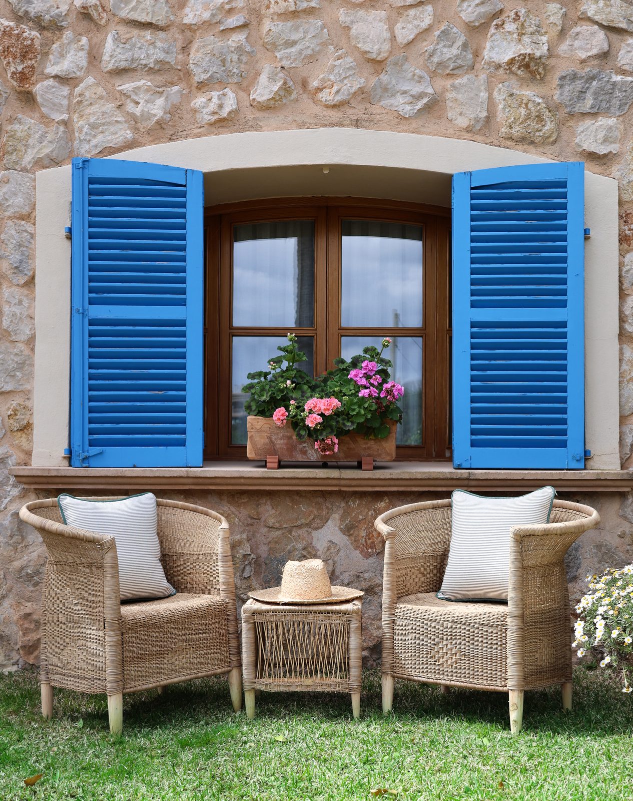 Traditional Mallorca window with blue louvred shutters, geraniums and two cane chairs on the lawn