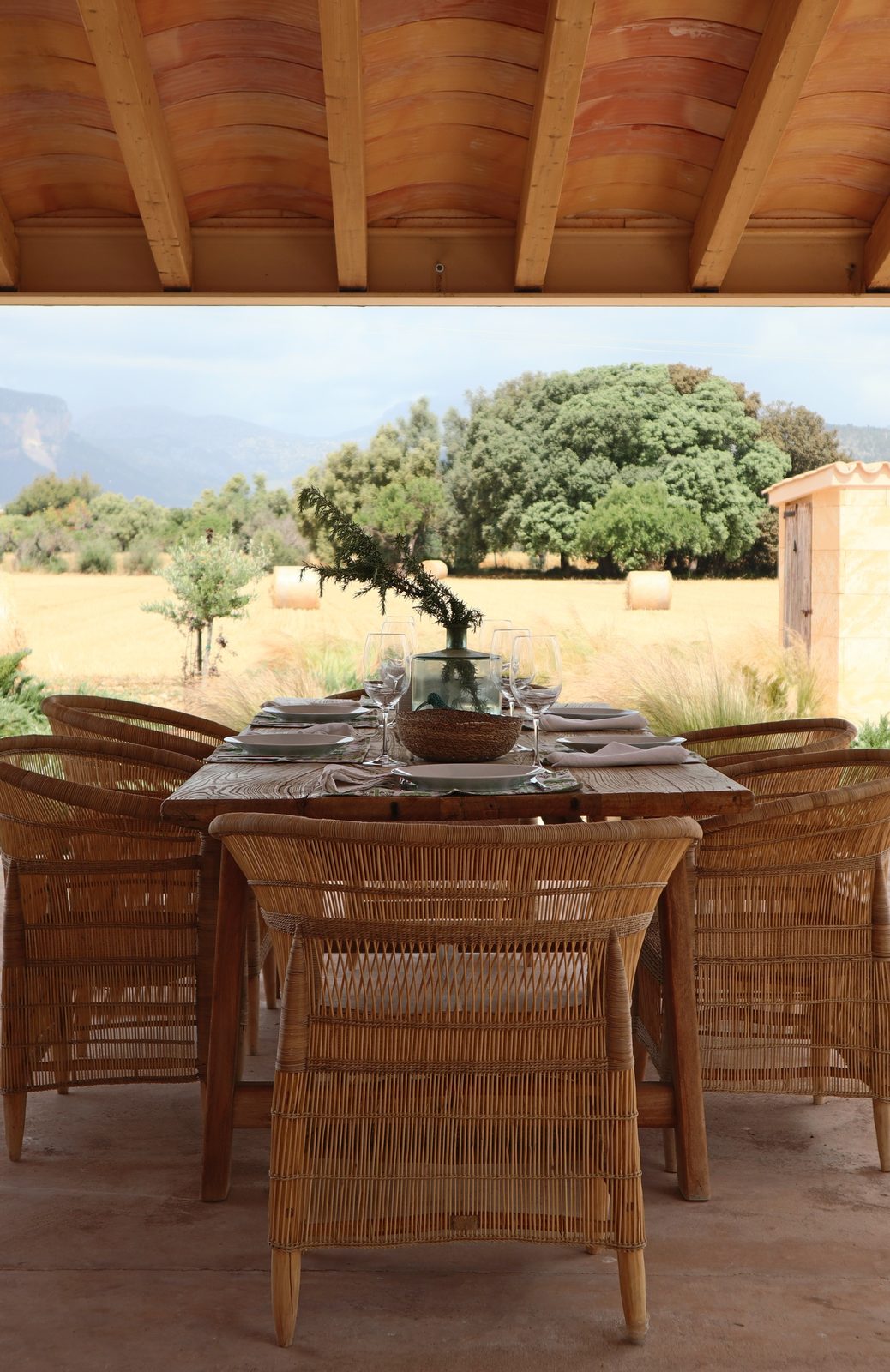 Covered terrace dining table set with woven chairs overlooking a hay field