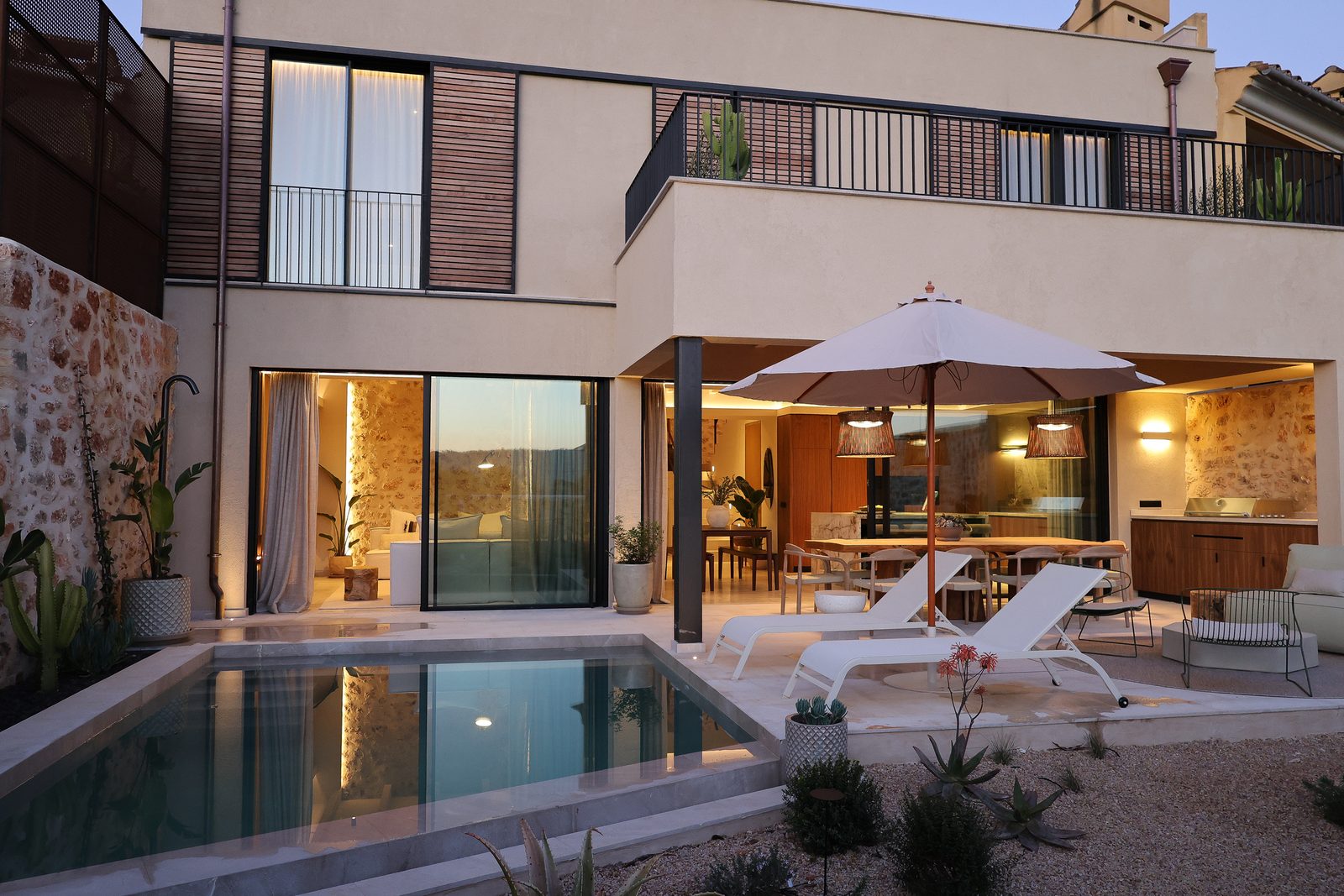 Contemporary Mallorca villa at dusk with a lit interior, pool, loungers and parasol